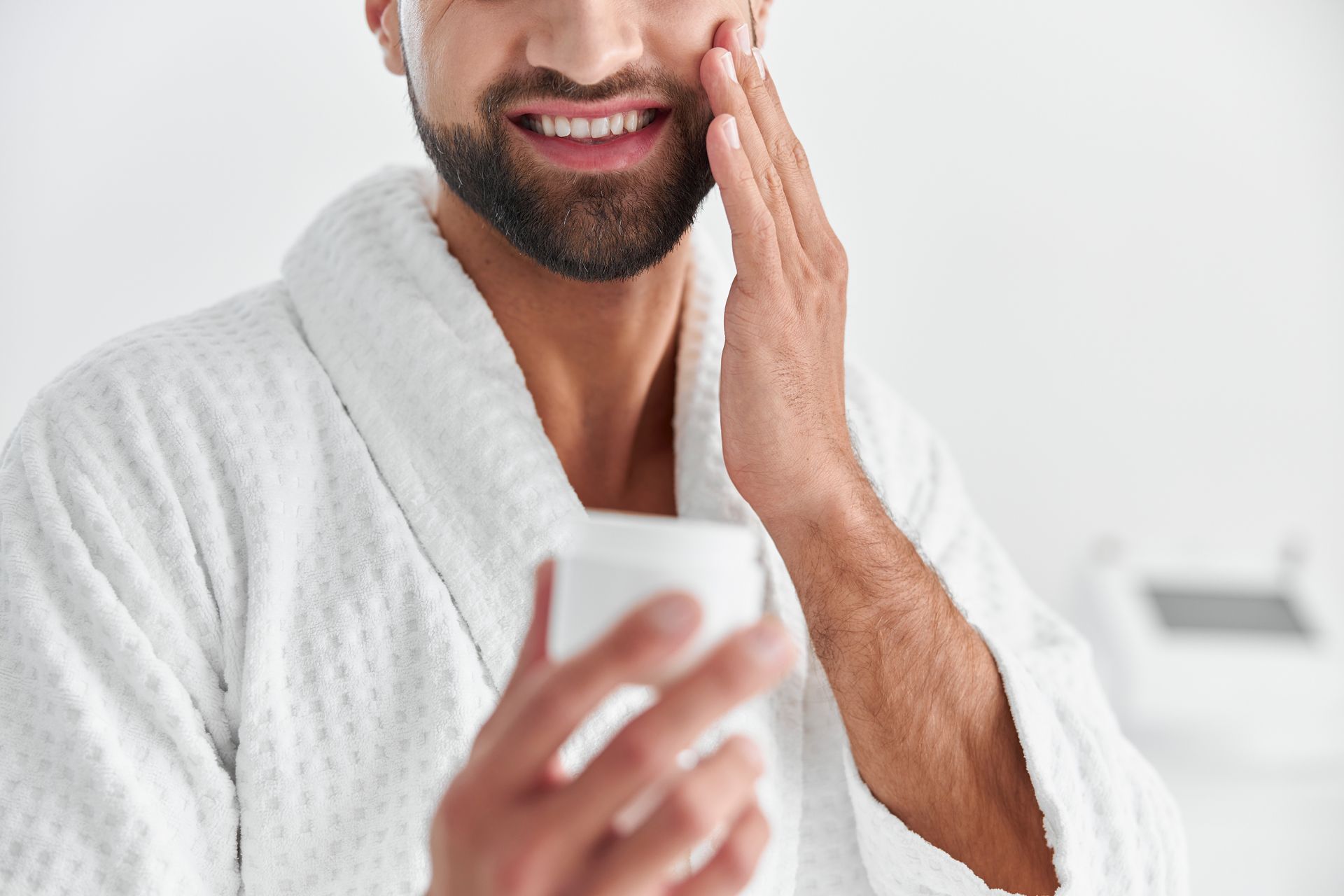 A man in a bathrobe is applying moisturizer on his face.