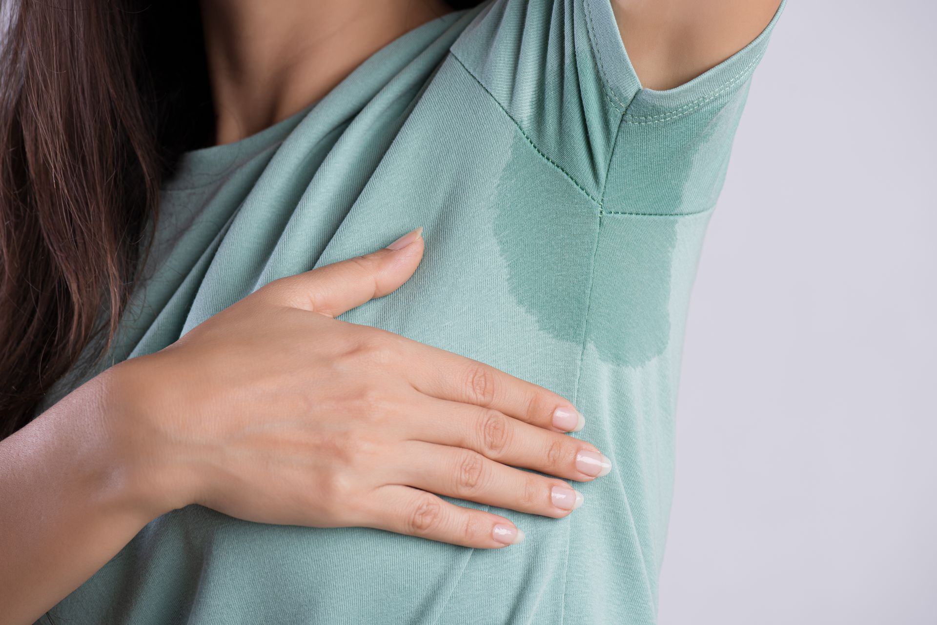 Woman touching damp armpit area of a light green shirt, sweat visible.