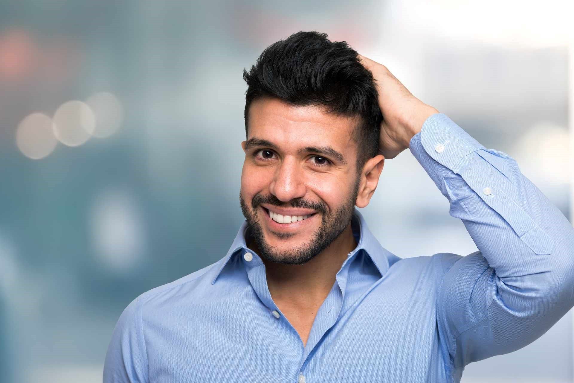 Smiling man in a blue shirt touching his hair against a blurred indoor background