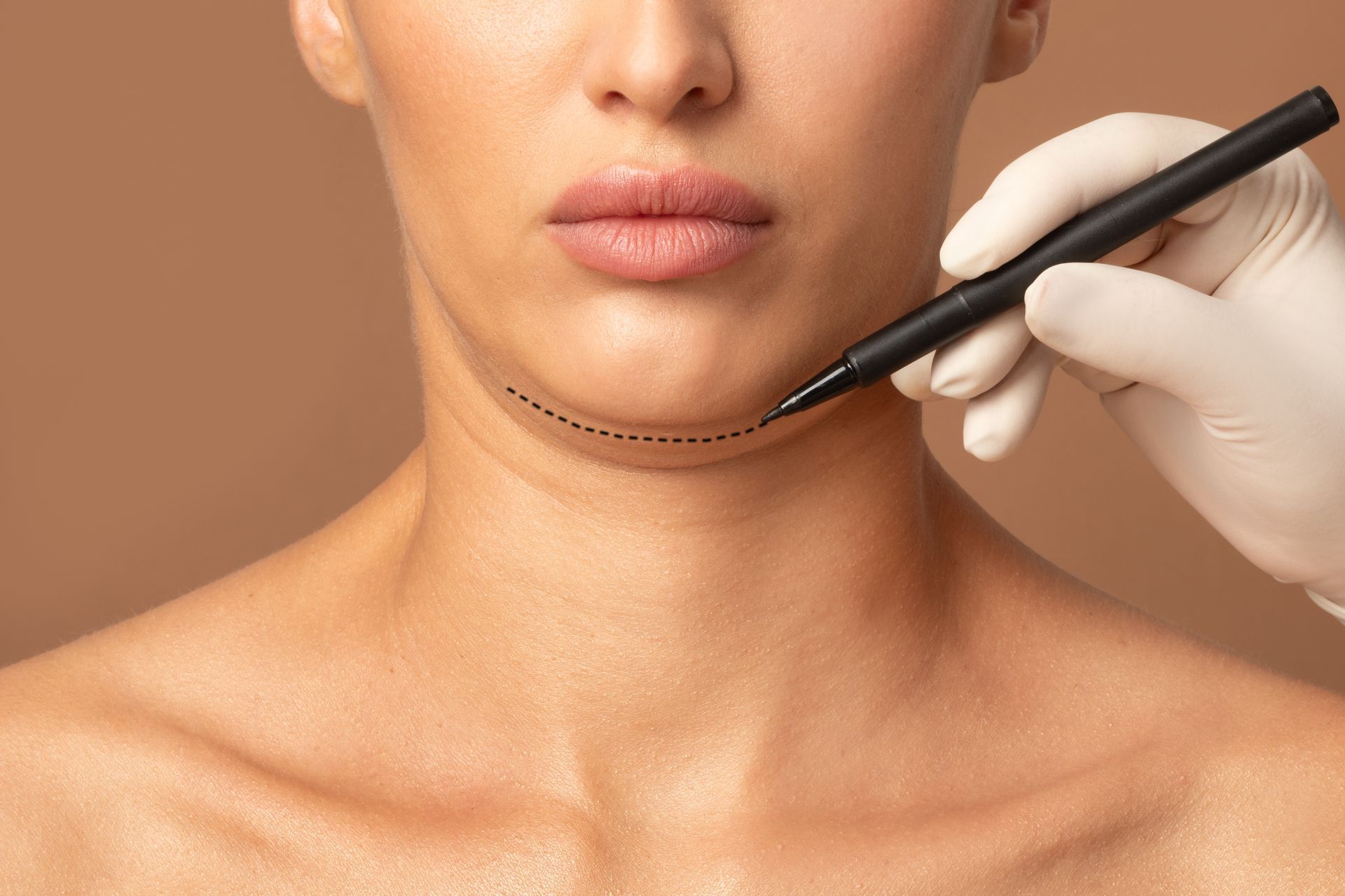 Gloved hand marks a jawline on a woman's neck with a black cosmetic pencil against a beige background