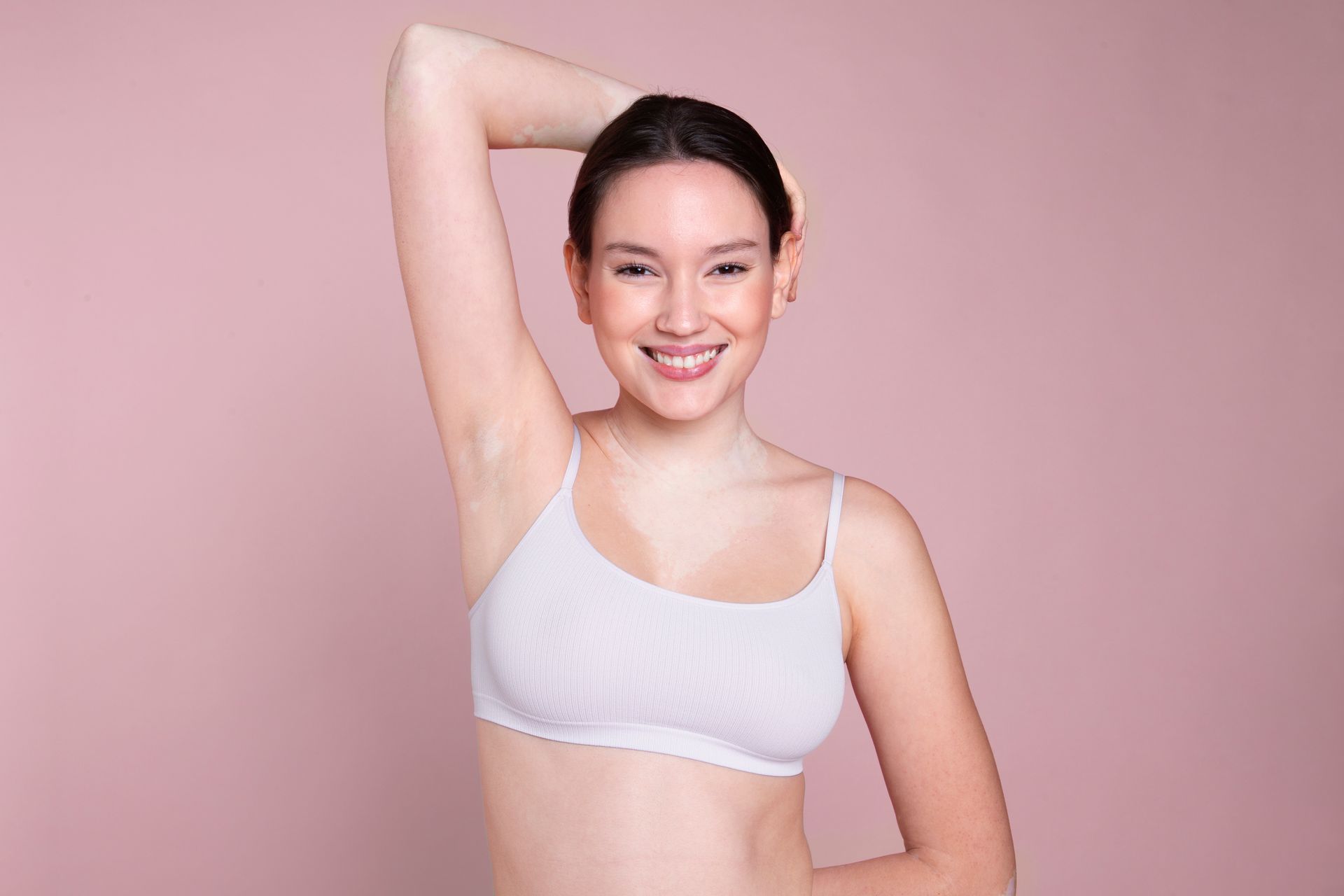 Woman in white bra, arm raised, smiling, against a pink background.