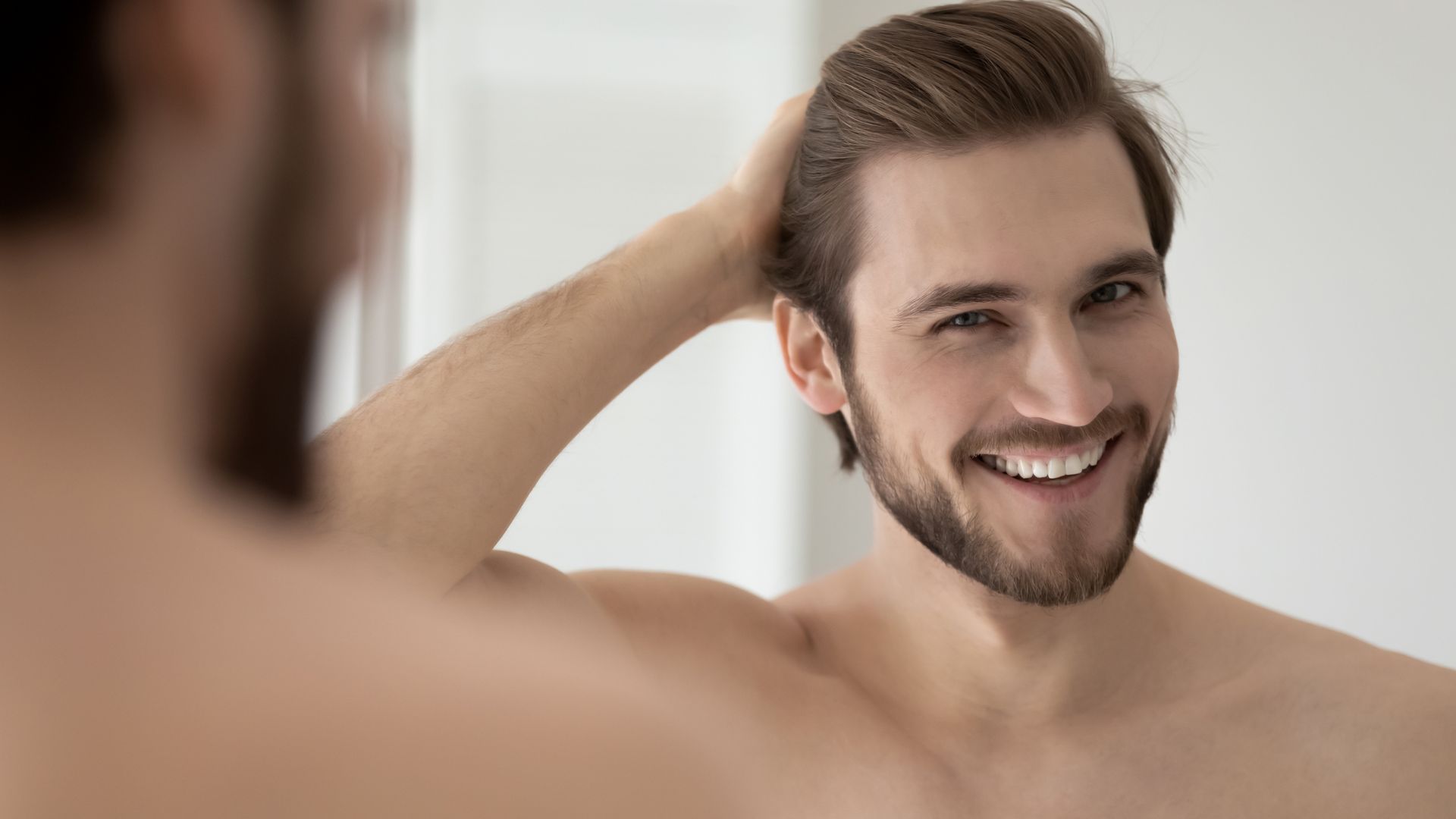 A shirtless man is smiling while looking at his hair in the mirror.