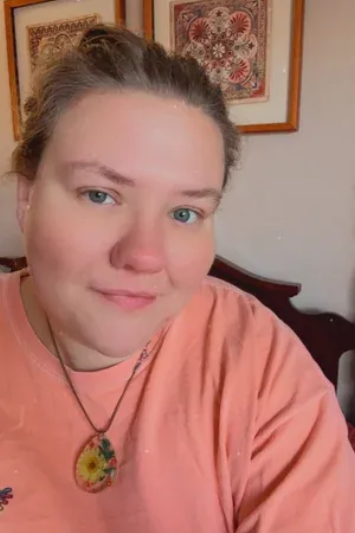 A person with light brown hair worn up, wearing a coral shirt and a floral pendant necklace, smiling indoors.