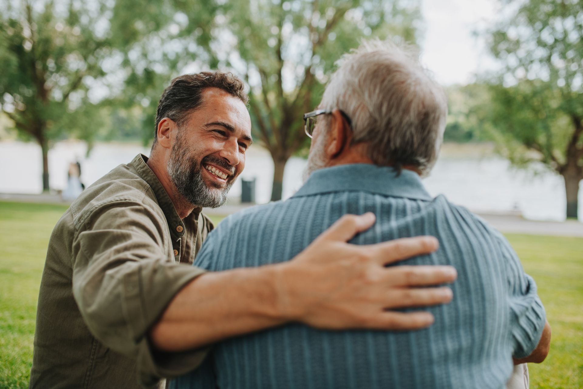 A person in a green shirt smiles while resting a hand on the shoulder of another person in a blue shirt outdoors.