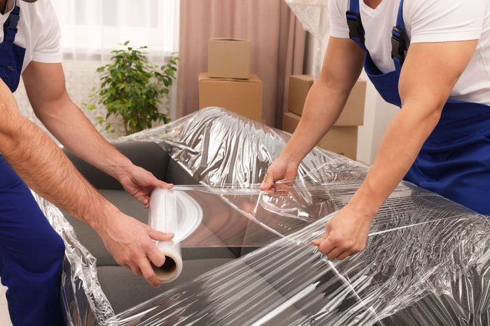 Two movers wrapping a gray couch in plastic wrap in a room with moving boxes.
