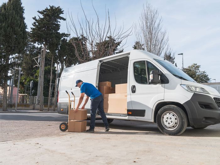 Delivery person loads boxes into a white van on a sunny day.