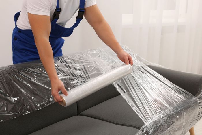 Person wrapping a gray couch with clear plastic wrap. White wall background.