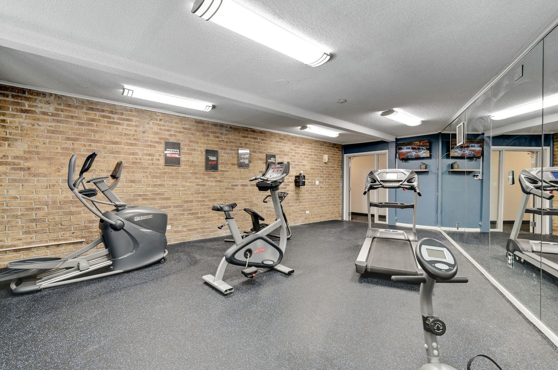 Gym interior with exercise equipment: elliptical, stationary bike, treadmill. Exposed brick wall and large mirror.