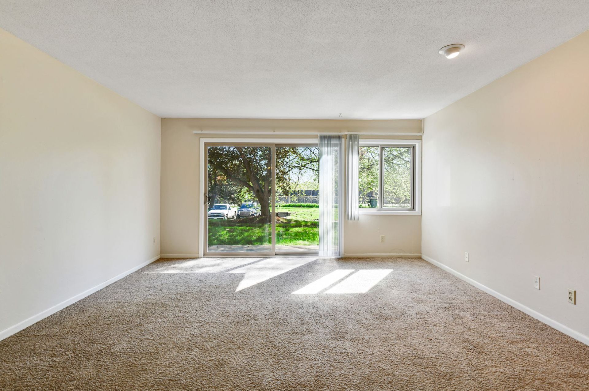 Empty living room with beige walls and carpet, large glass door to a park view at Manor Royal in Plymouth, MN.