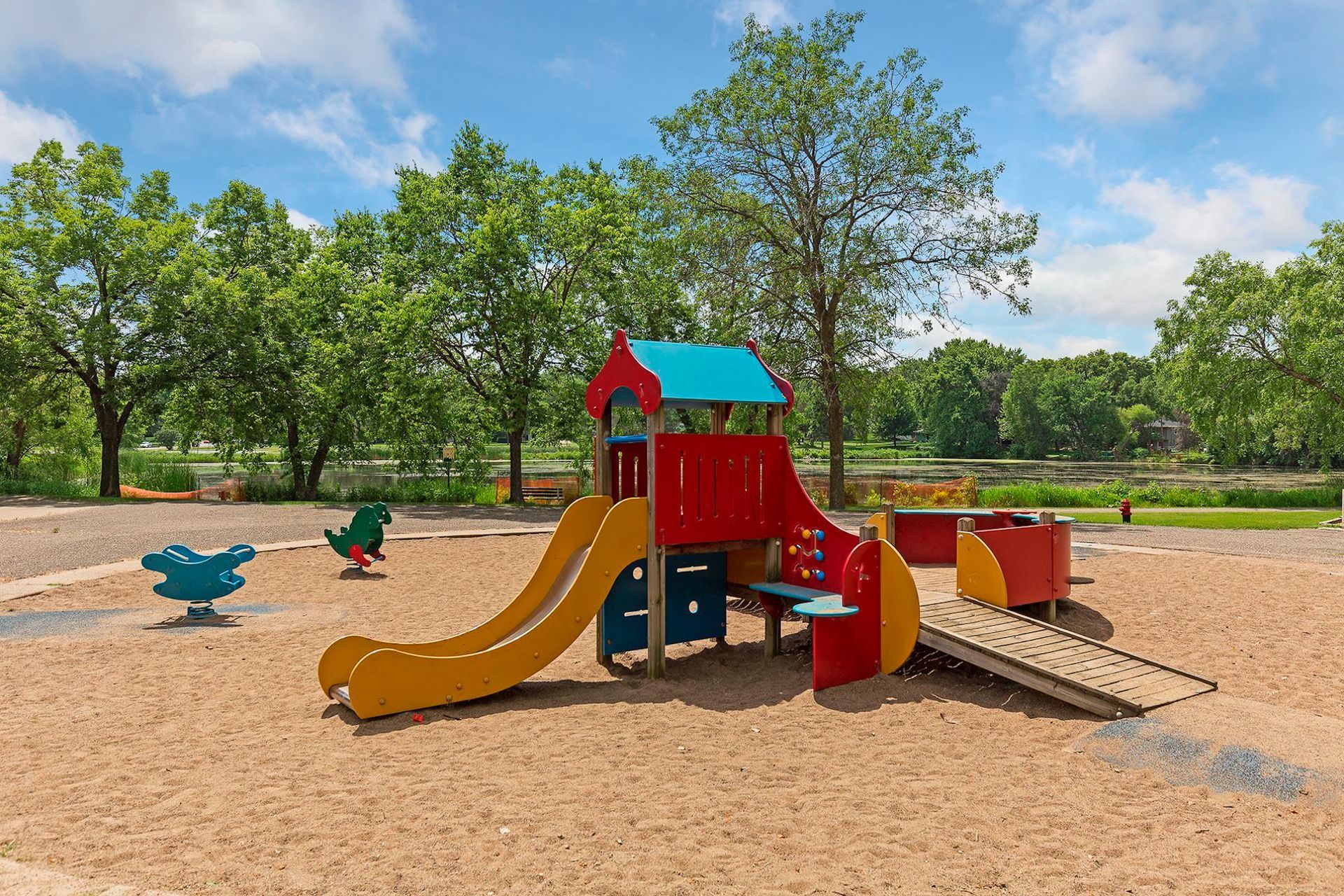 Playground with slide and bridge, surrounded by trees and a blue sky at Manor Royal in Plymouth, MN.