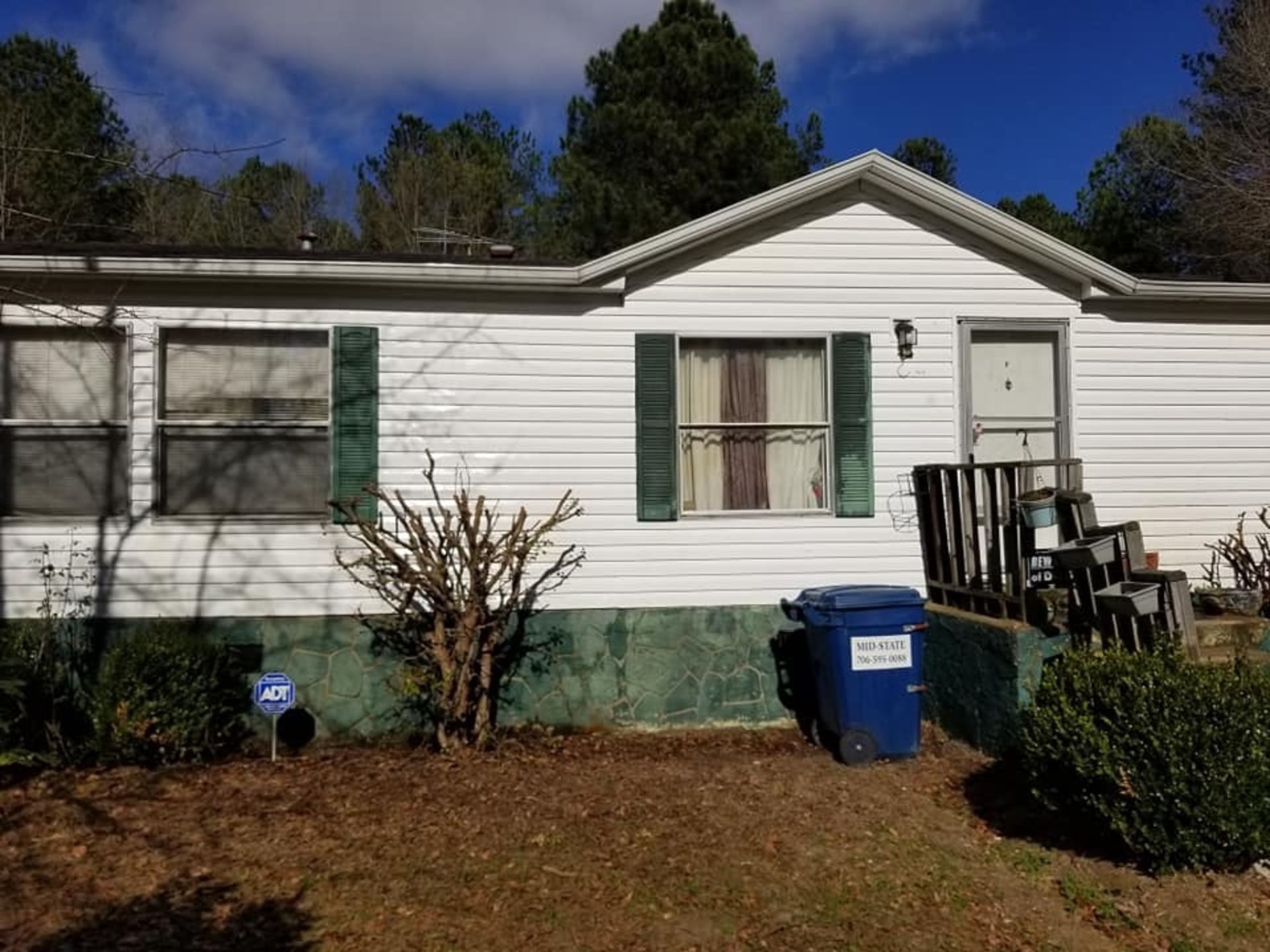 A white mobile home with green shutters and a blue trash can in front of it.