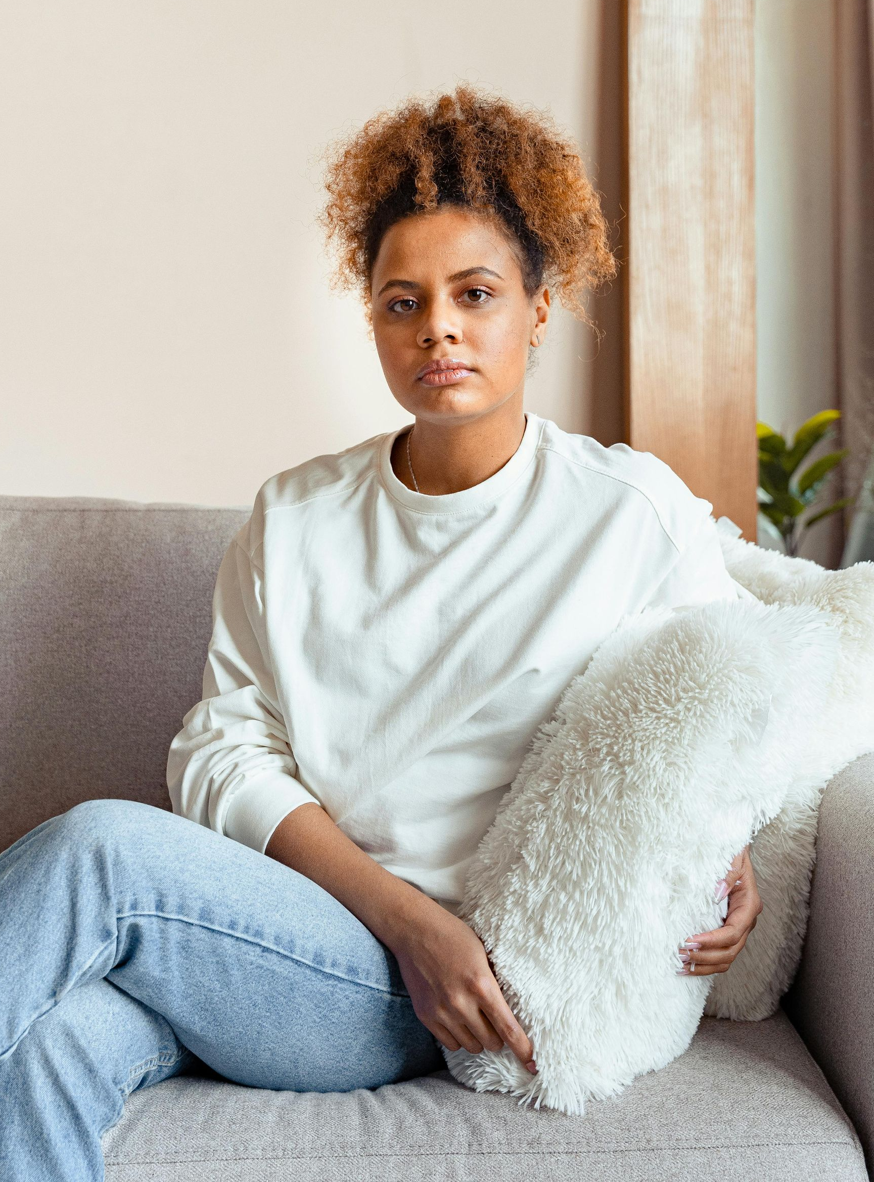 Woman with curly hair sits on couch, holding a fluffy white pillow. Wearing a white sweater and jeans. Indoors.