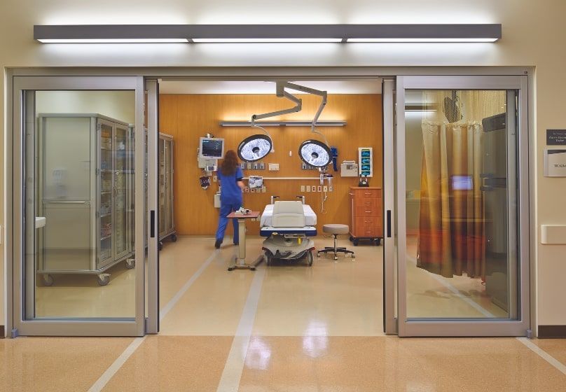 Sliding glass doors reveal an operating room with a person in scrubs preparing for a procedure.