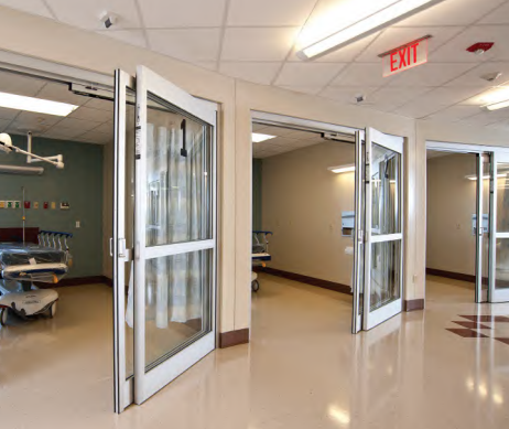 Hospital hallway with open glass doors, medical equipment visible in rooms, and 