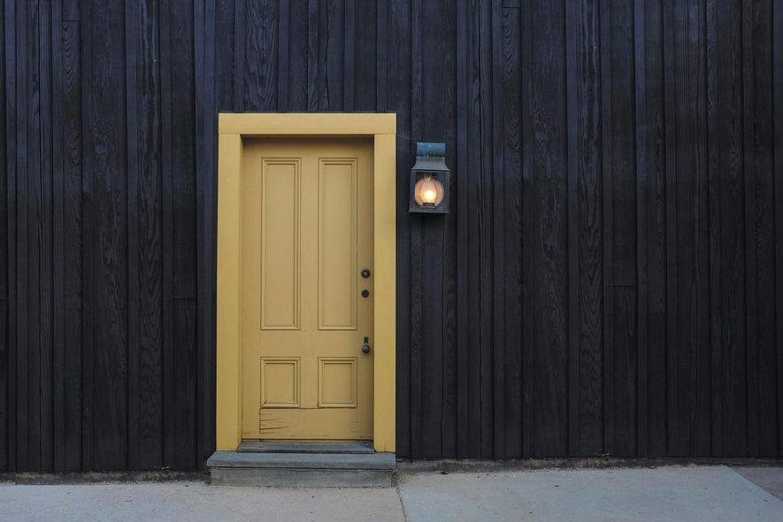 Yellow door in a yellow frame set in dark wood siding, a lit lantern to its right.