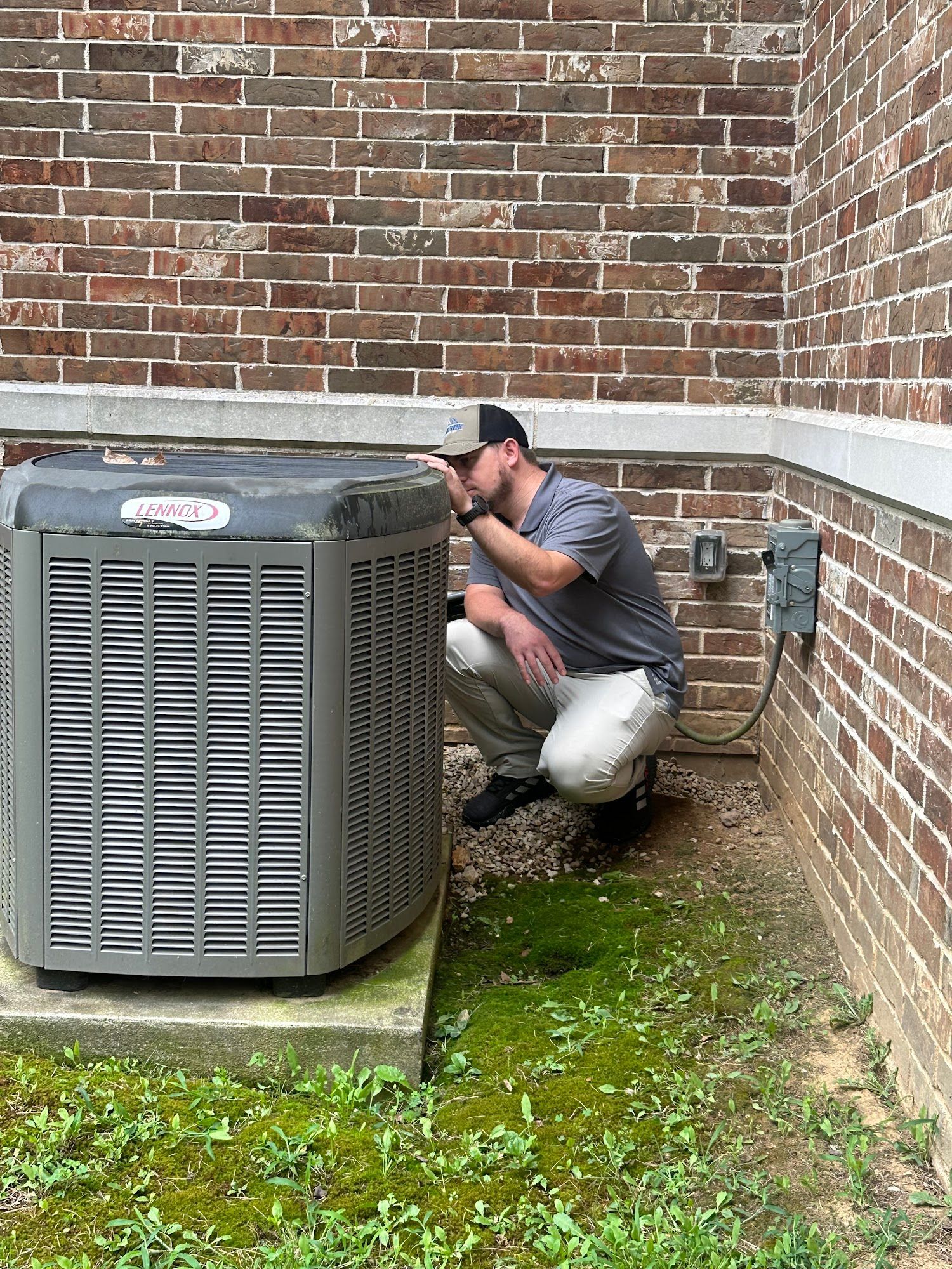 Man inspecting a gray air conditioning unit next to a brick wall. He's squatting, looking closely.