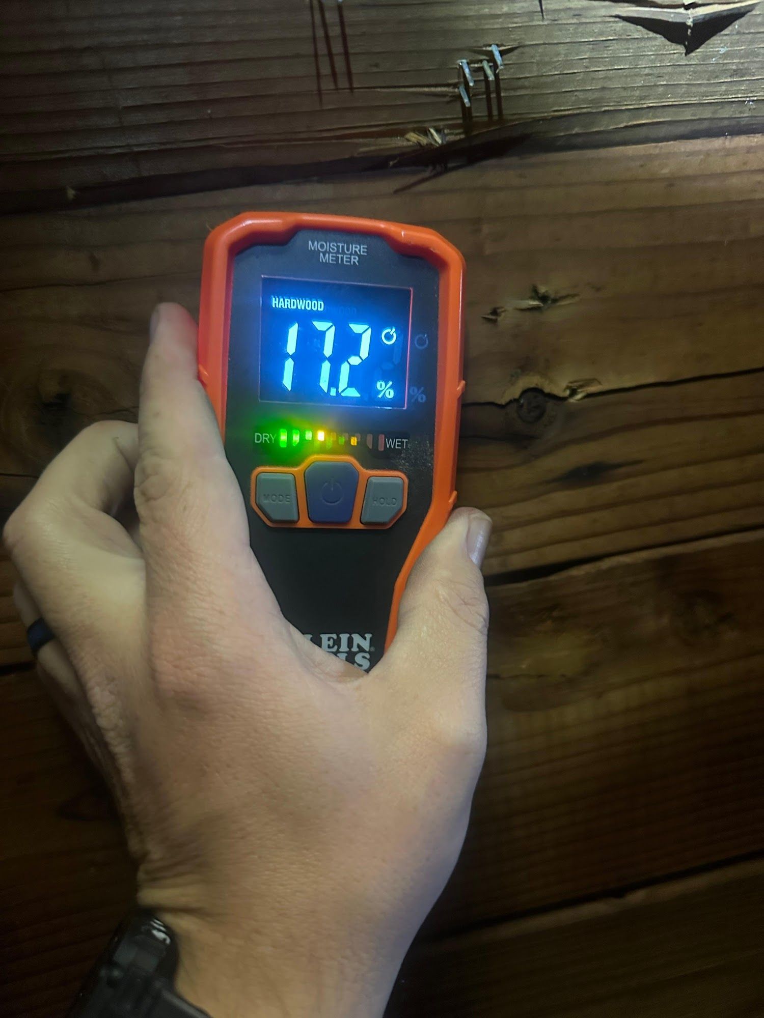 Hand holding a Klein Tools moisture meter against wooden boards; display reads 17.2°C and 7%.