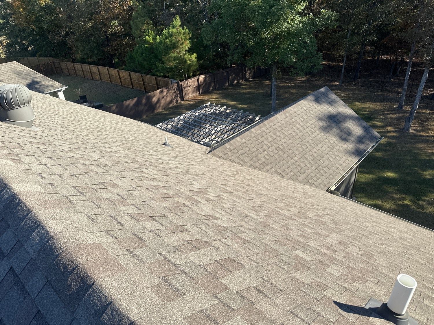Brown shingle roof with a chimney and vent. Trees and a fence in the background.