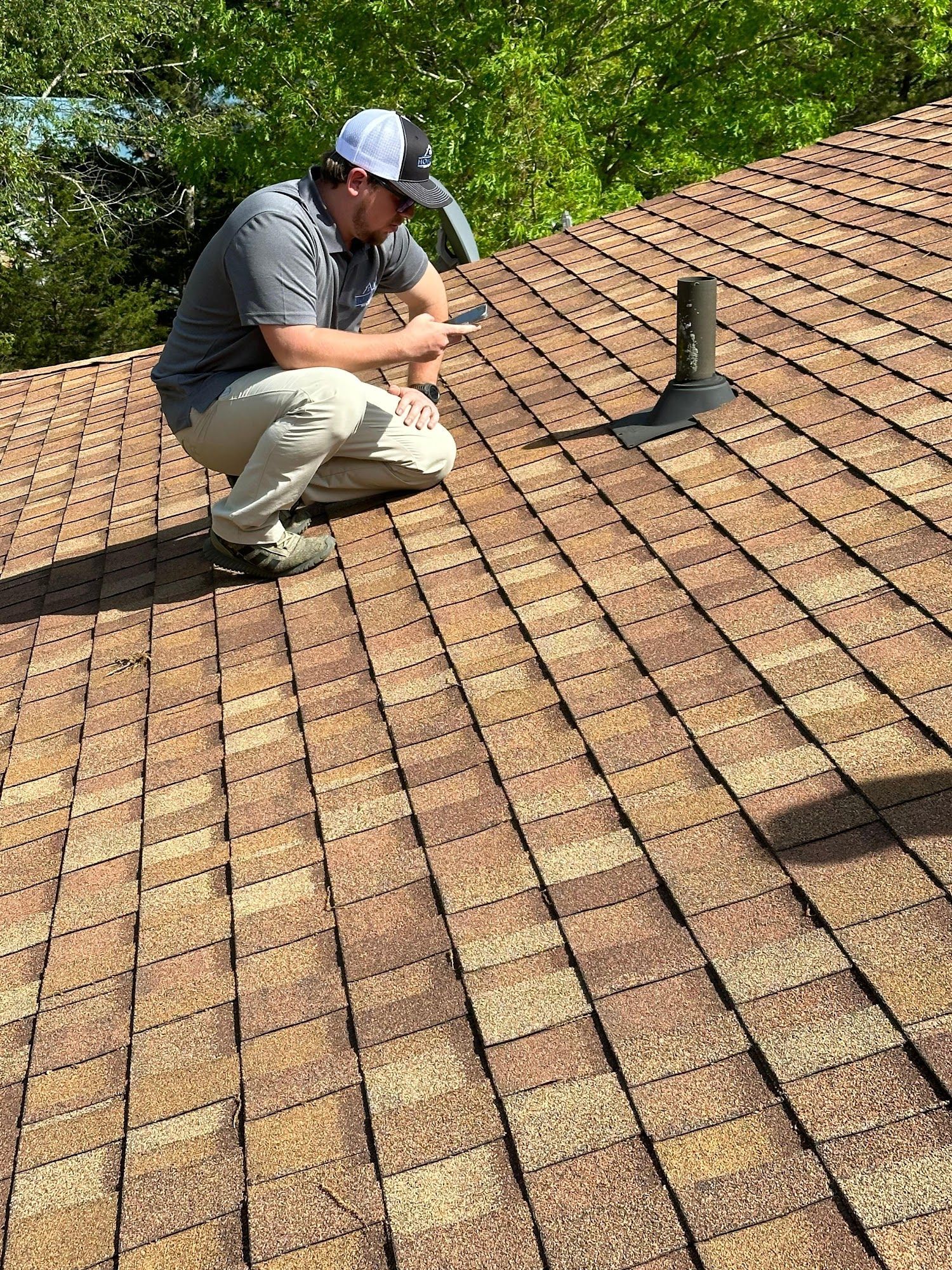 Man on roof inspecting shingles near a vent pipe; brown and tan roof tiles, sunny outdoors.