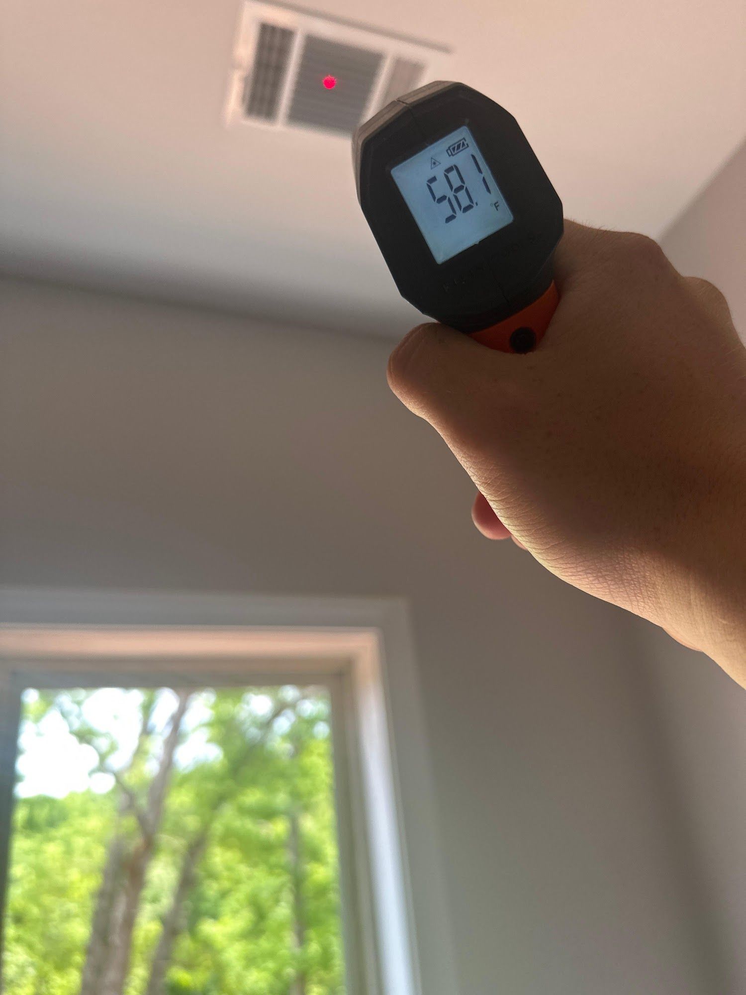 Hand holding thermometer pointed at ceiling vent, reading 58.1. Bright room with window.