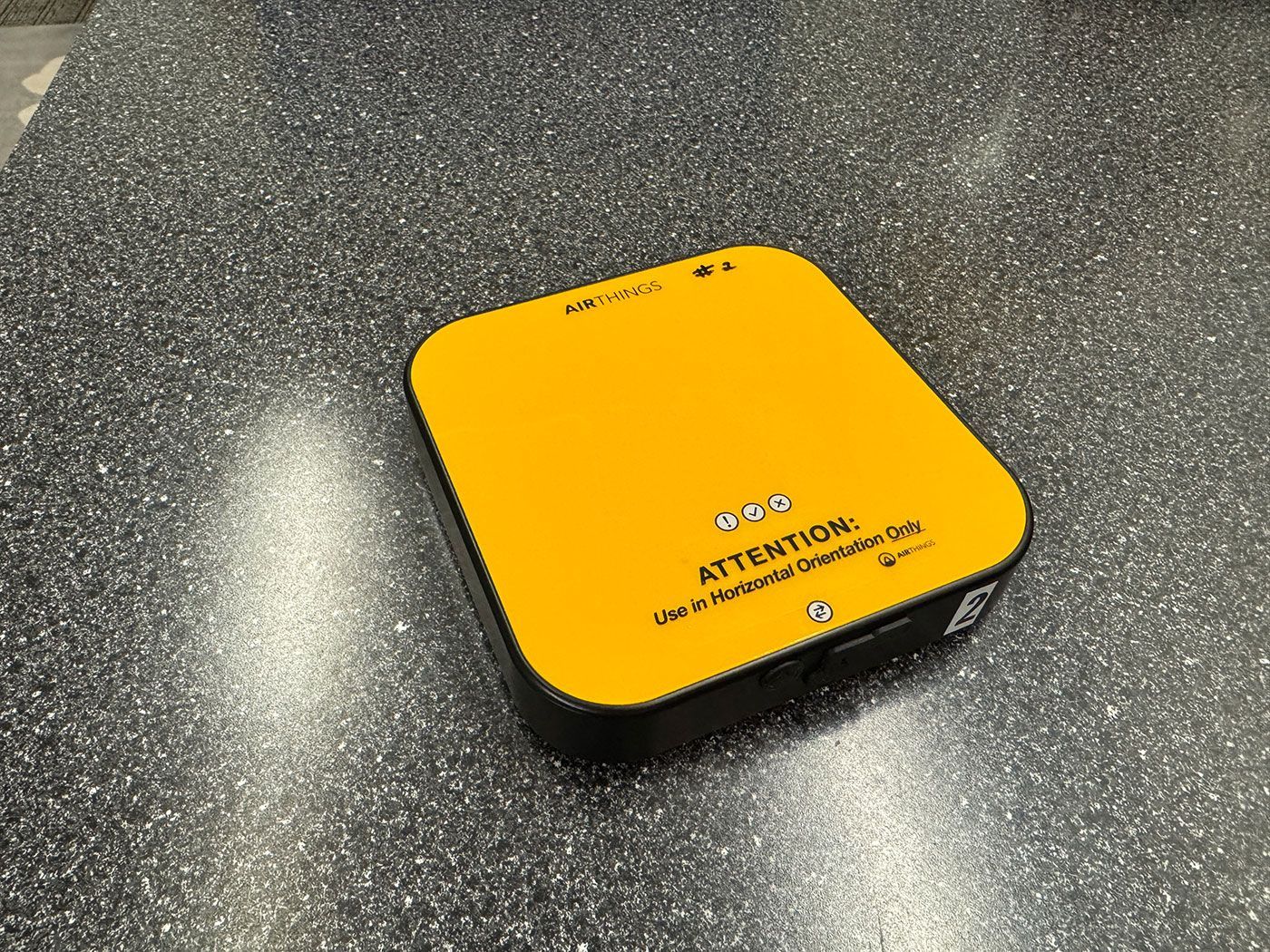Yellow and black square device on a textured gray surface; it has text: