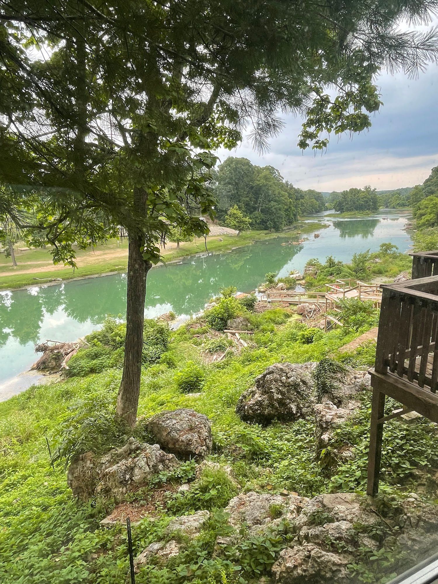 A river winds through a green landscape with trees and large rocks, viewed from a deck.
