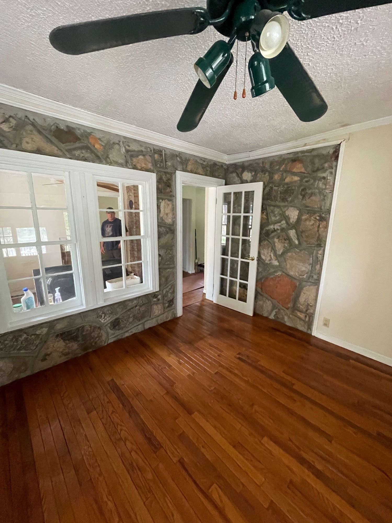 Room with stone walls, wood floor, white windows, and french doors; a dark green ceiling fan is overhead.