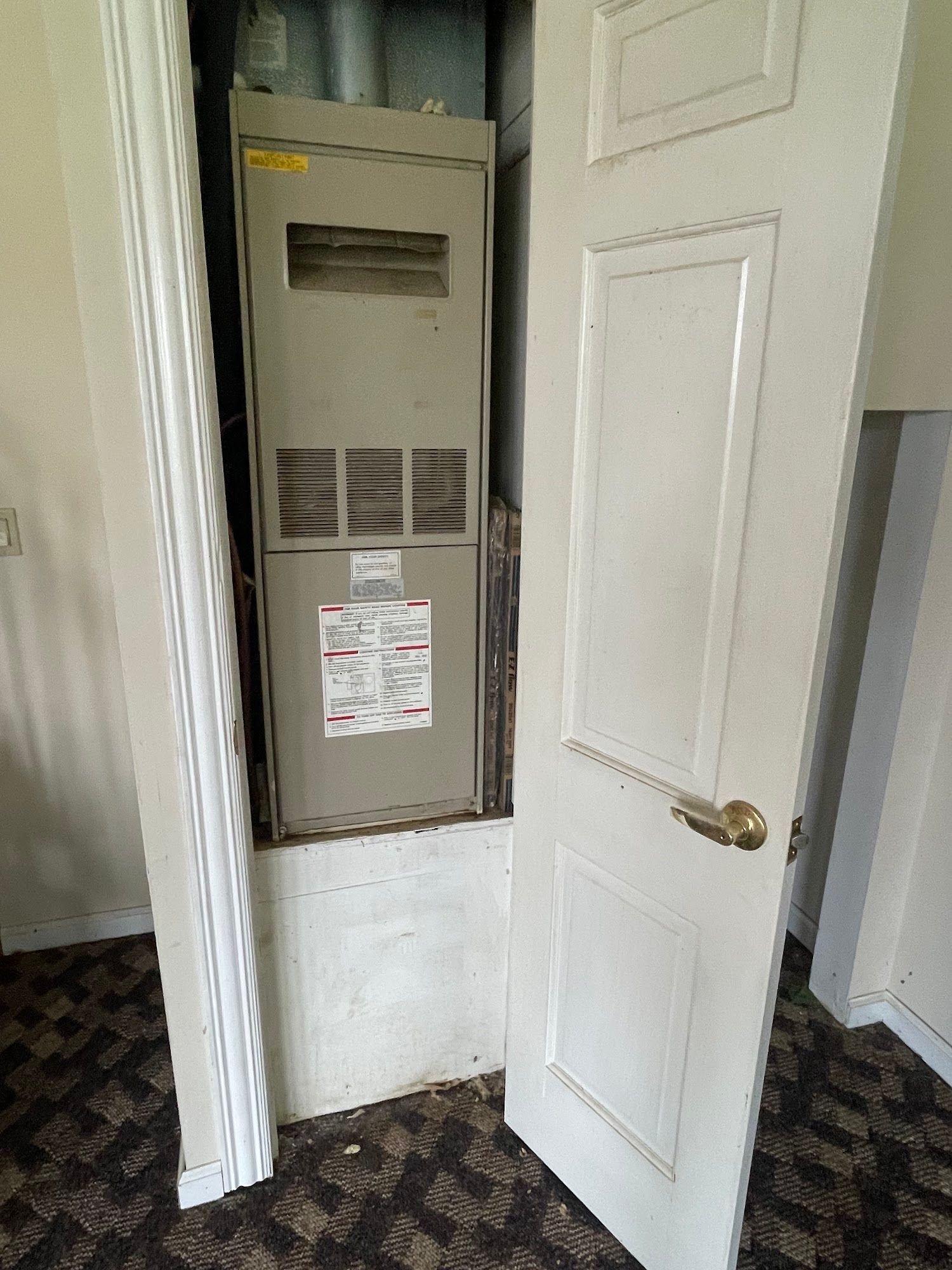 Furnace unit inside a closet, door open. Gray metal and white door and trim. Dark patterned flooring.