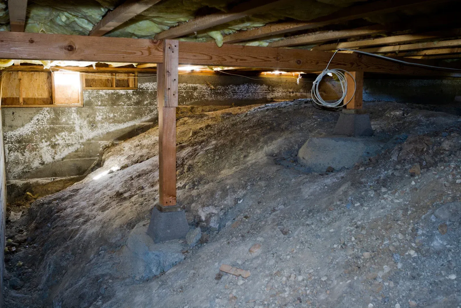 Crawlspace with wooden beams, posts, and dirt/gravel floor. Insulation visible above.