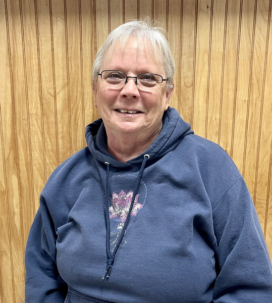 A woman wearing glasses and a blue hoodie is smiling in front of a wooden wall.