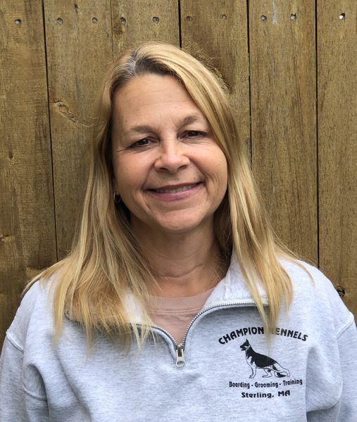 A woman wearing a gray champion kennel sweatshirt is smiling in front of a wooden fence.