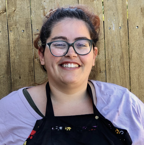 A woman wearing glasses and an apron is smiling in front of a wooden fence.