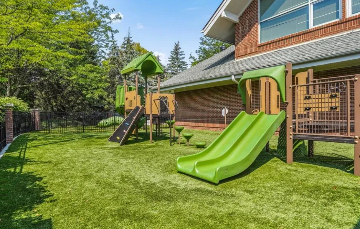 A backyard with a green playground set and a swing set on Artificial Turf.