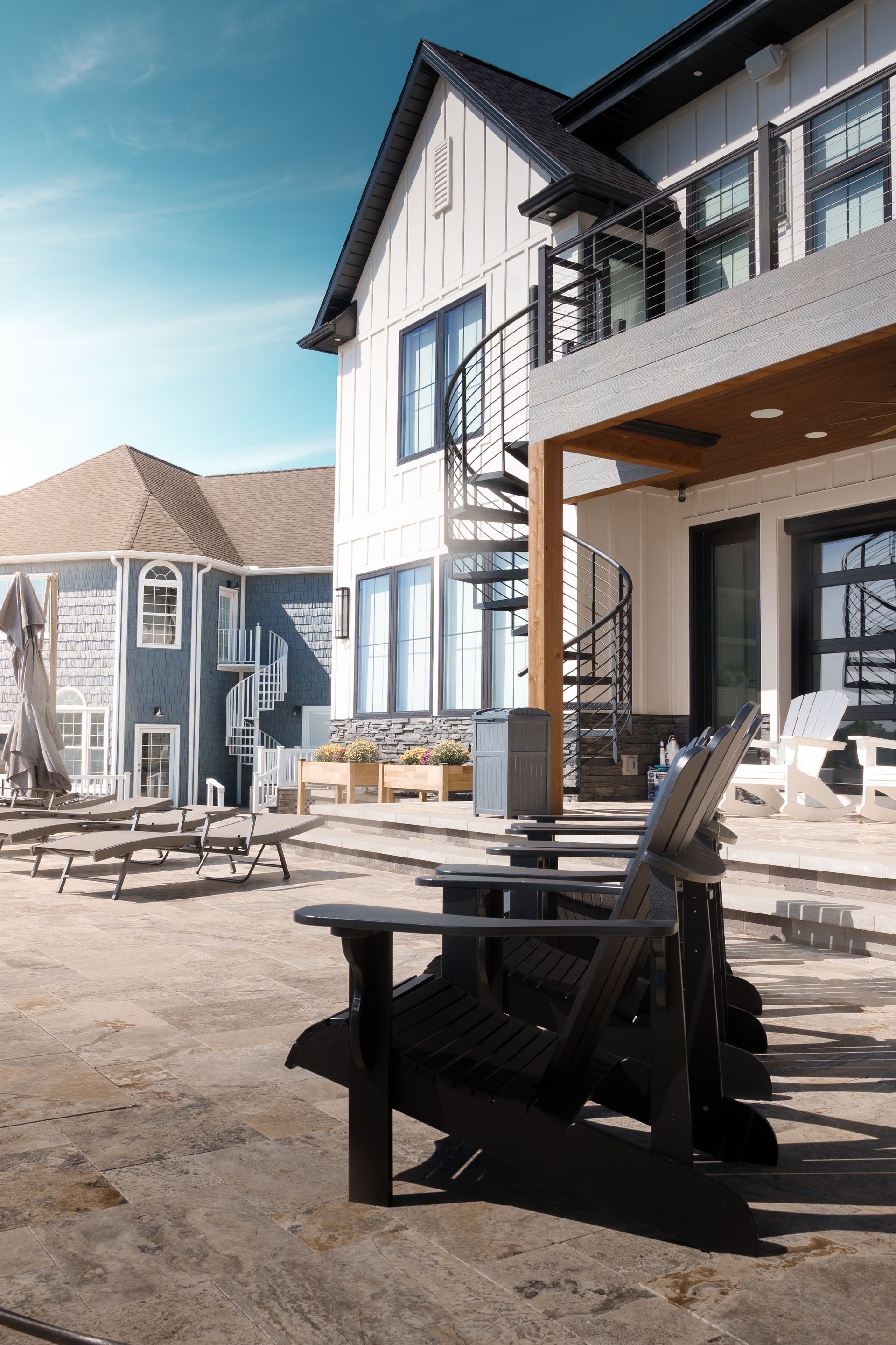 A modern white building with a spiral staircase and a row of black Adirondack chairs faces a sand courtyard. A blue building is visible in the background.