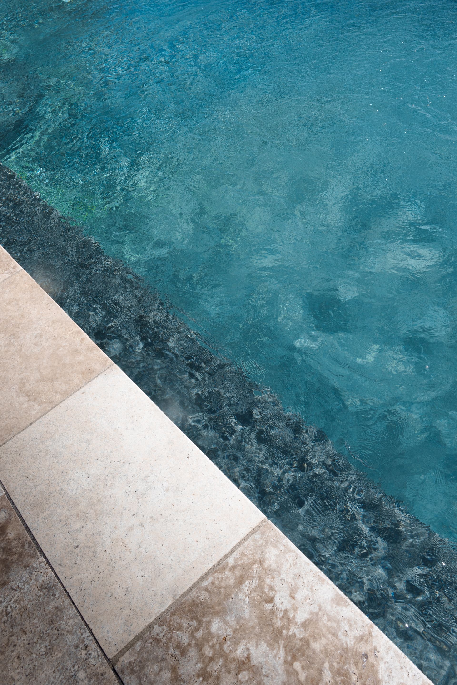 Edge of a pool: Beige stone tiles meet a dark speckled border next to clear turquoise water.