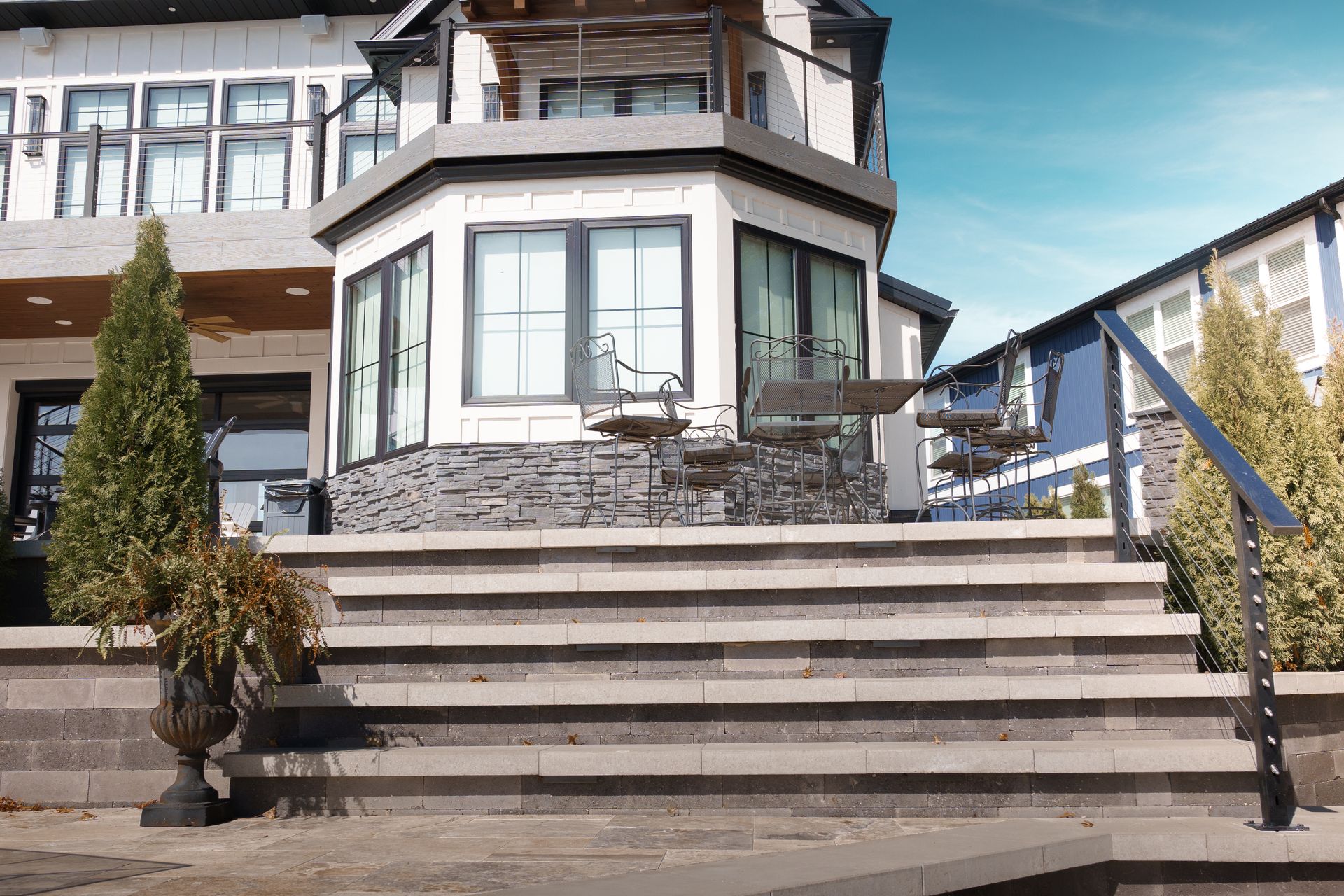 A two-story house with stone steps and a black metal handrail. A small tree and potted plants decorate the exterior.