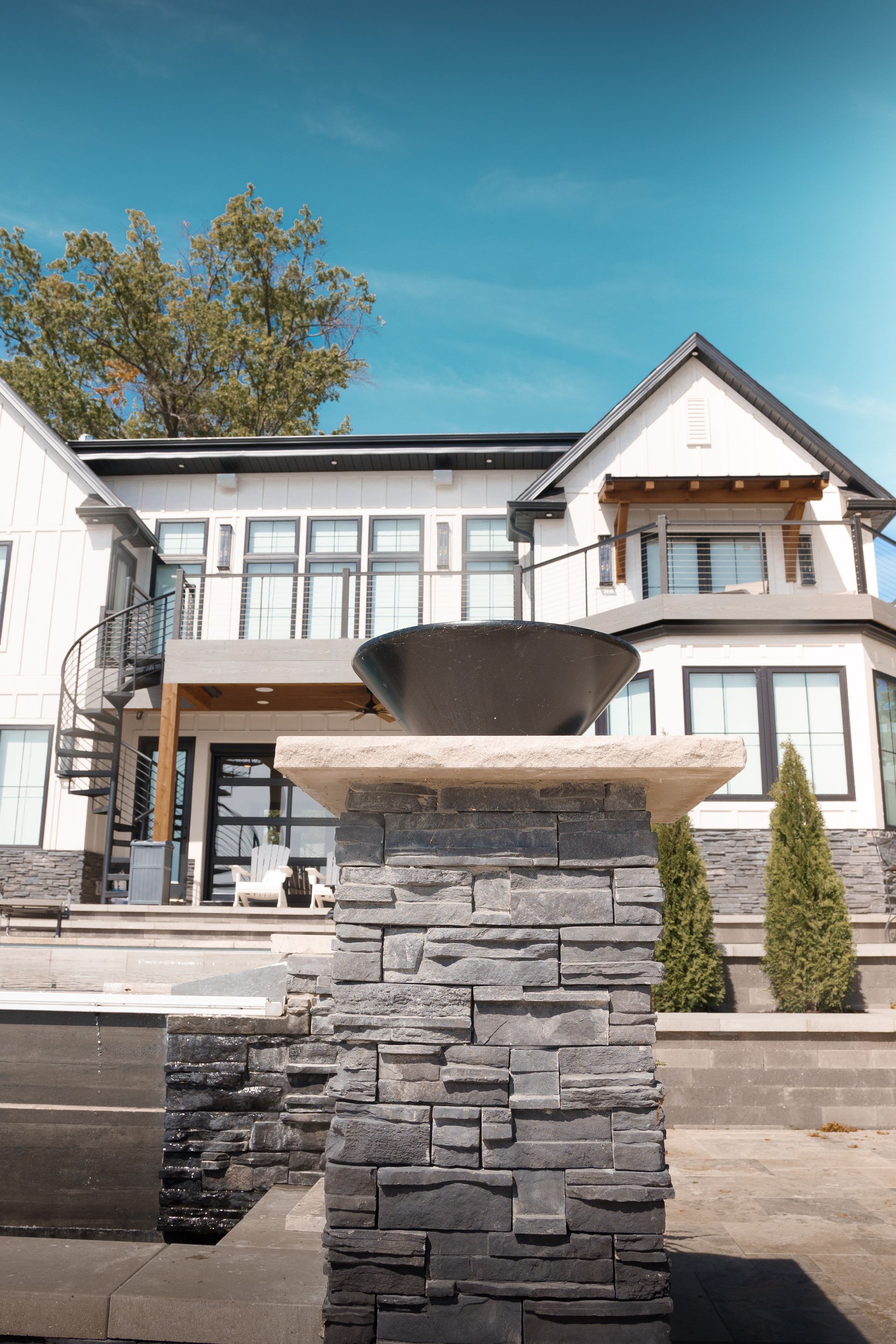Stone pillar with a dark bowl, in front of a modern house with a balcony and spiral staircase against a blue sky.