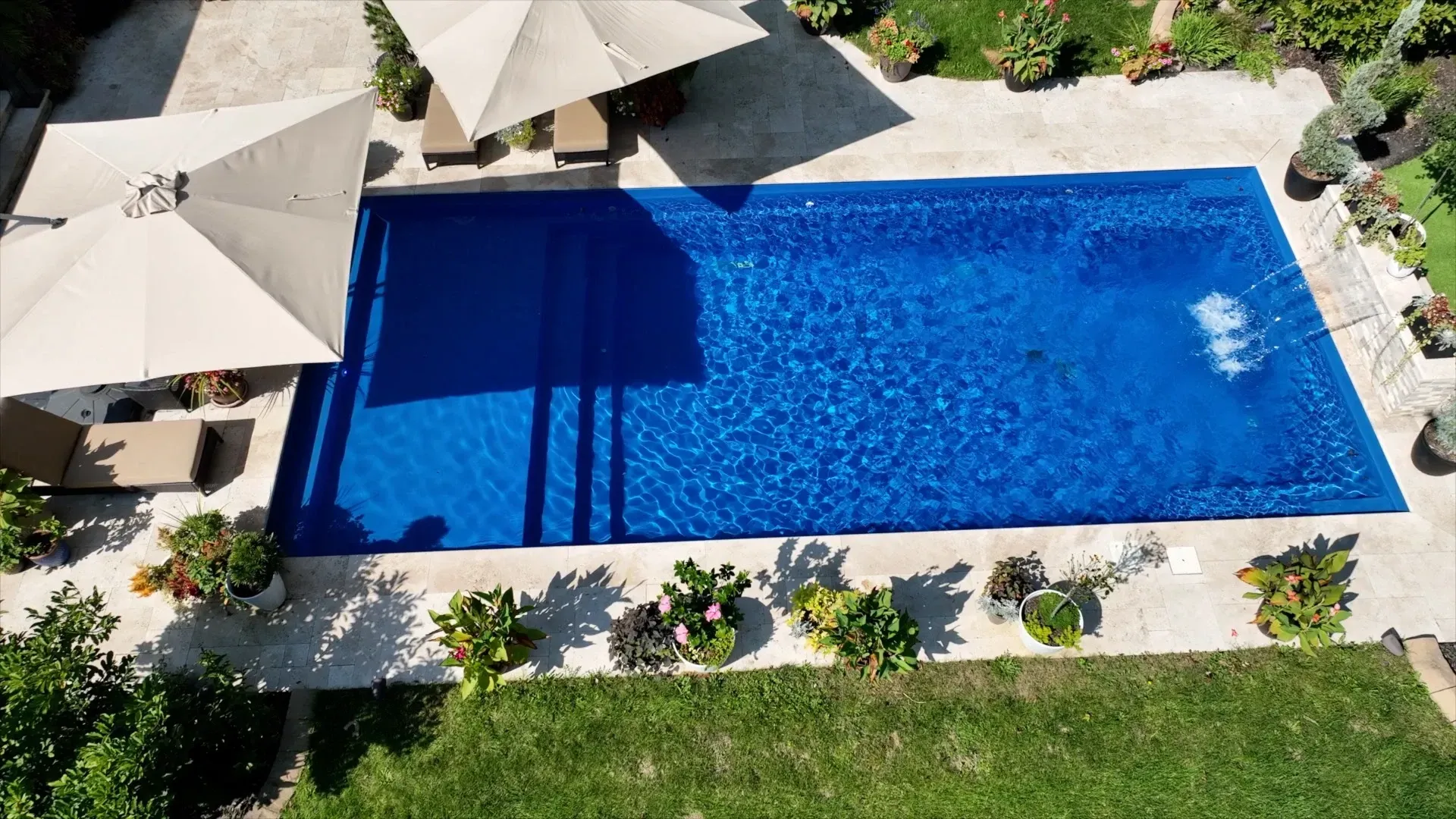 Overhead view of a rectangular swimming pool with blue water, flanked by stone patio, greenery, and two open umbrellas.