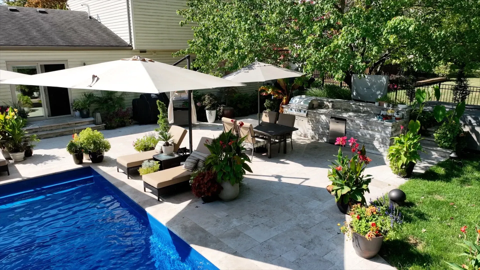 Backyard patio with pool, lounge chairs under umbrellas, outdoor kitchen, and lush landscaping.