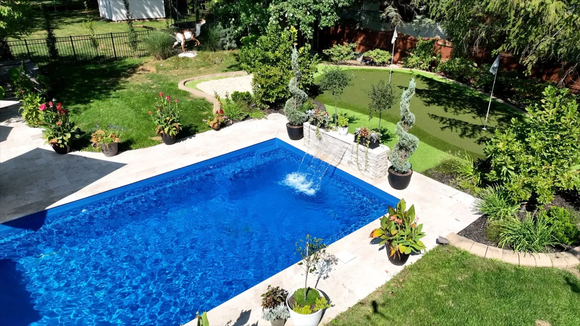 A blue rectangular pool surrounded by a patio, lush greenery, and potted plants in a backyard setting.