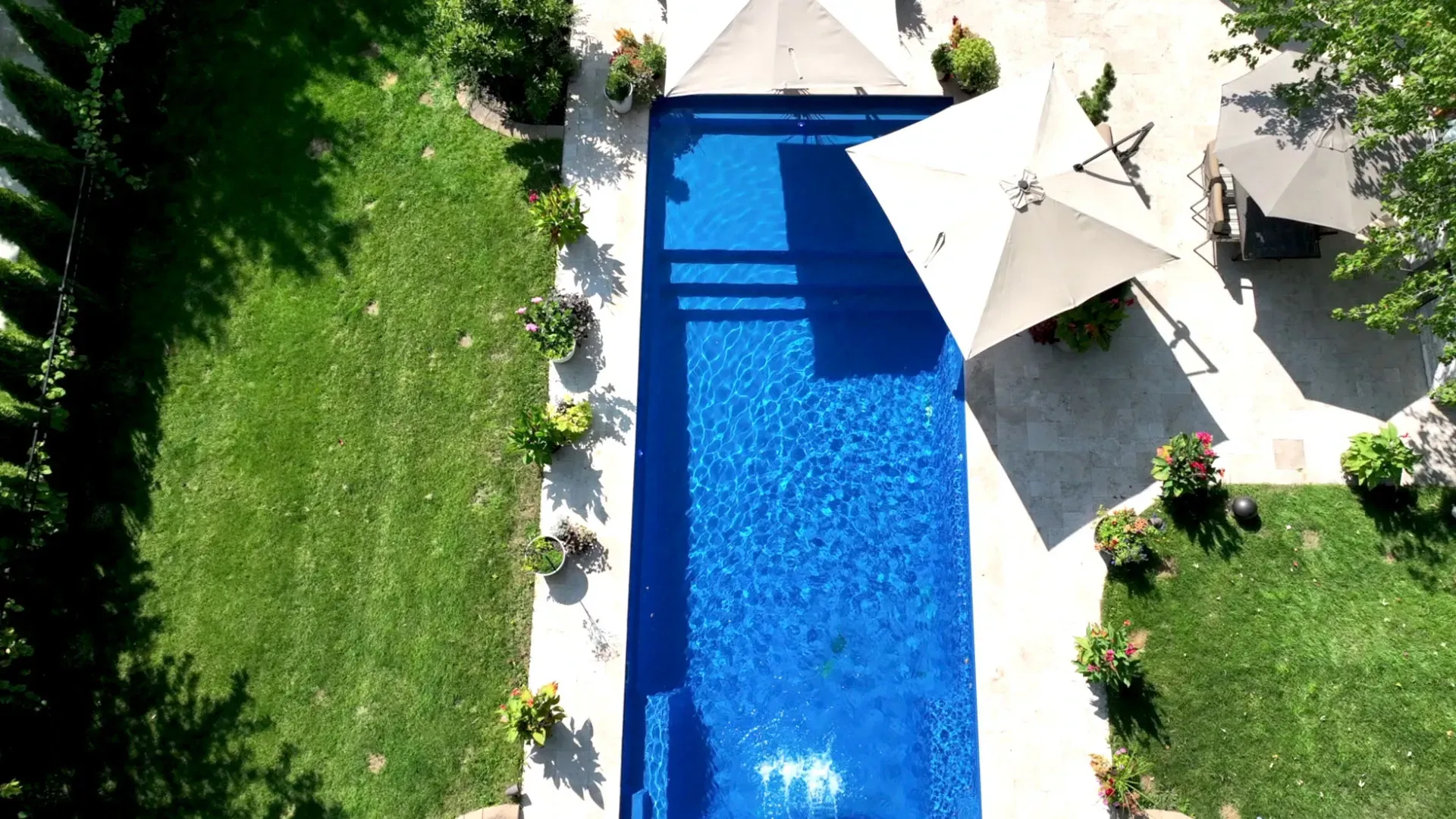 Aerial view of a rectangular blue swimming pool surrounded by green grass and white concrete. Two large umbrellas provide shade.