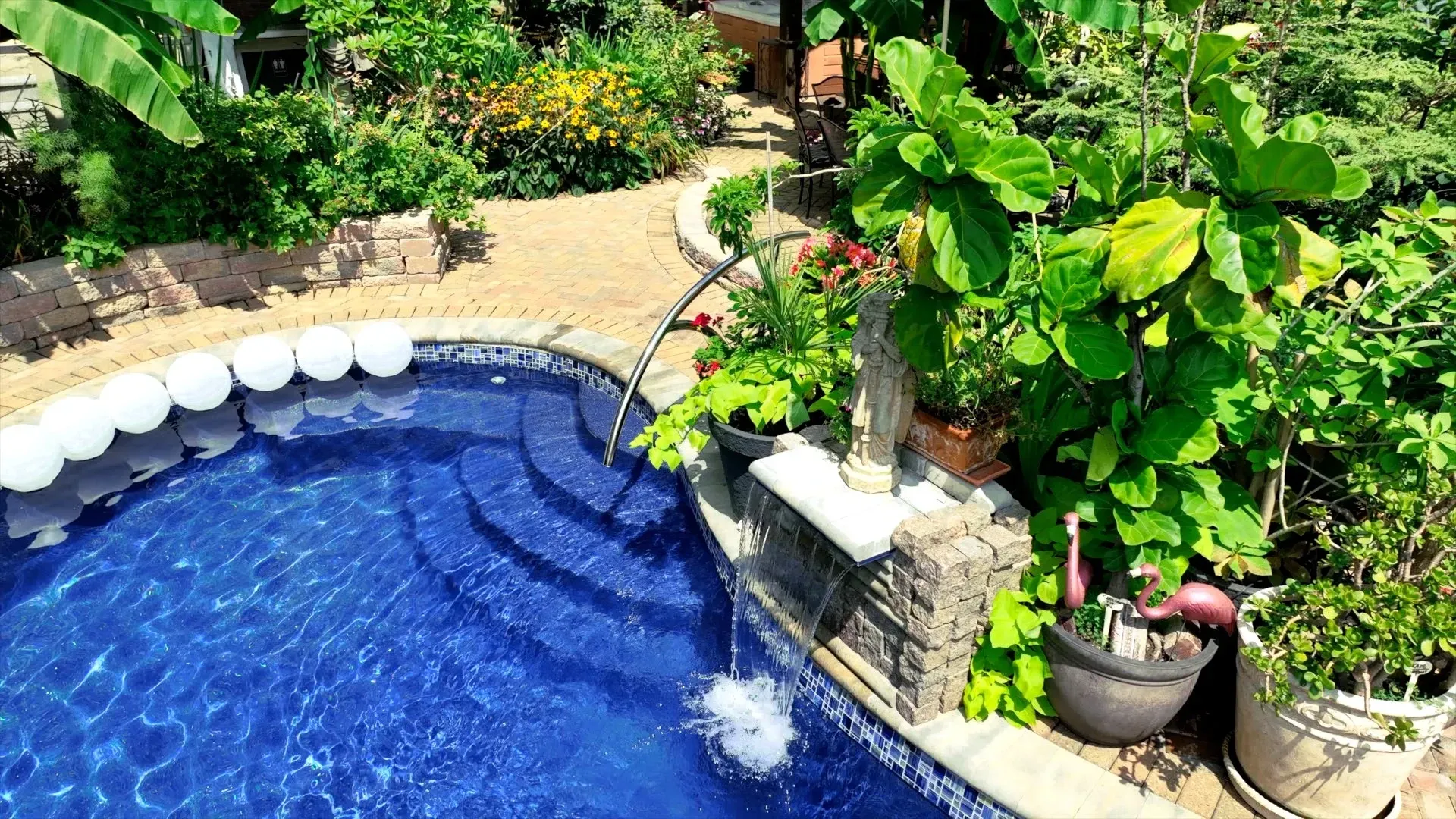 A blue-tiled pool with steps, surrounded by greenery, a stone wall, and a fountain, in a sunny outdoor setting.