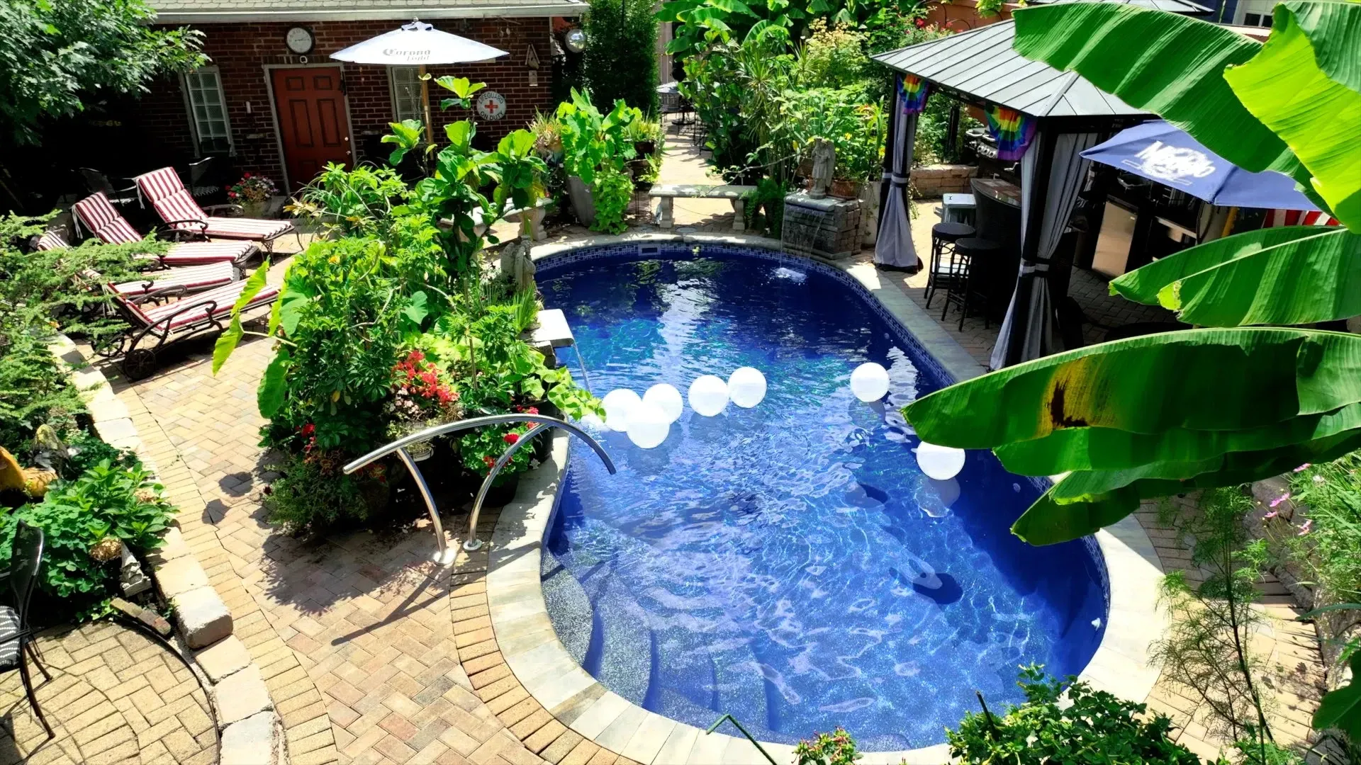 Backyard oasis with a pool, lounge chairs, a gazebo, and lush greenery. White balloons float in the blue water.