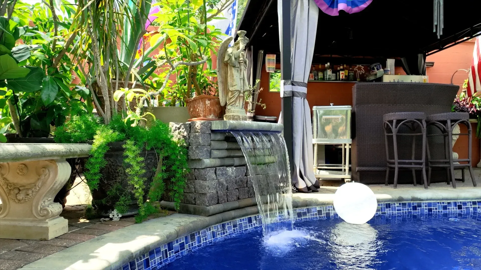 A backyard pool with a water feature, bar, and seating. A glowing orb floats in the blue water.