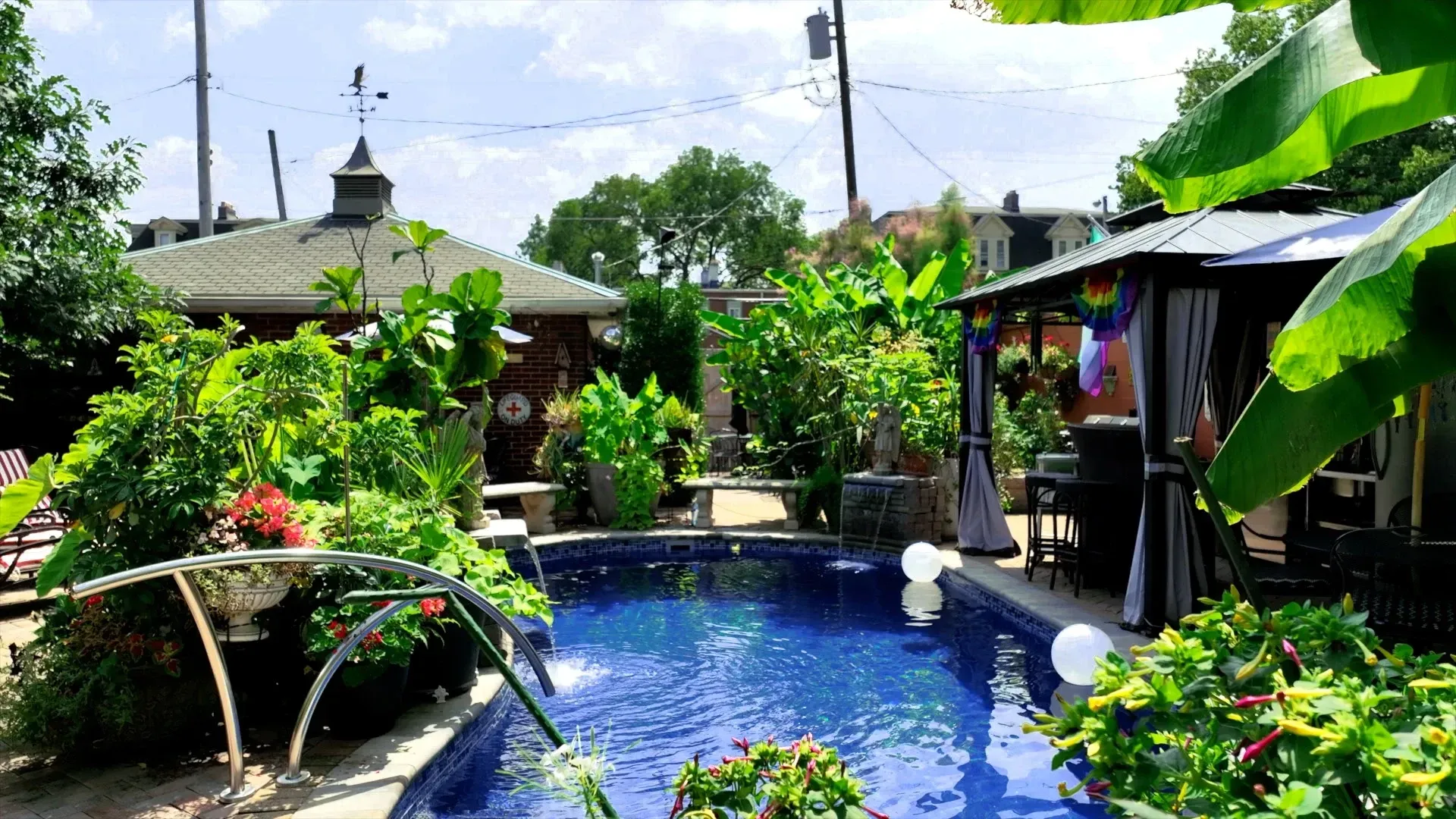 A lush backyard with a pool and gazebo. Green foliage surrounds the pool, and a building with a weathervane is in the background.
