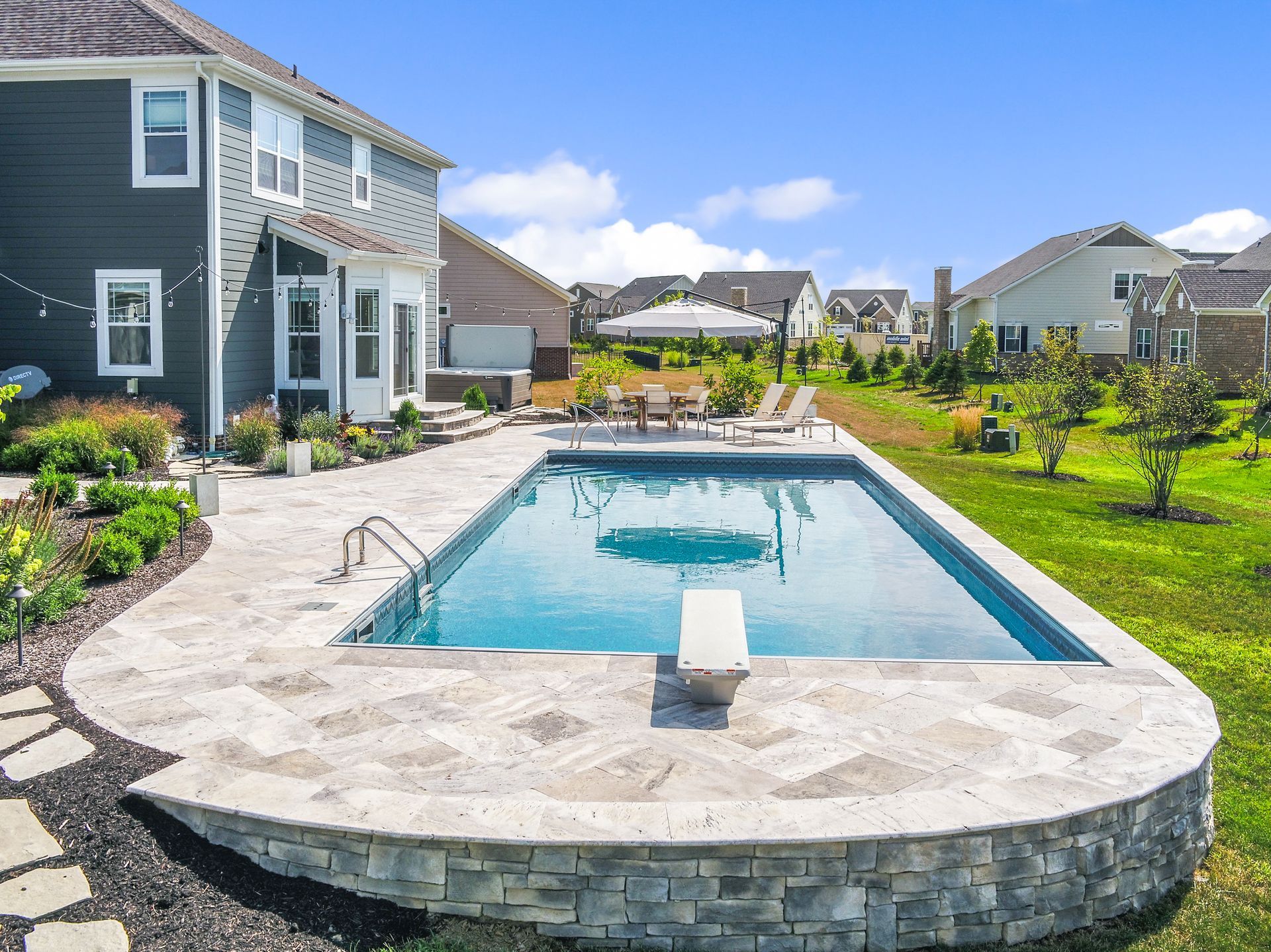 Backyard with a rectangular pool, diving board, and stone patio. Blue water reflects a bright sky, surrounded by green grass and houses.