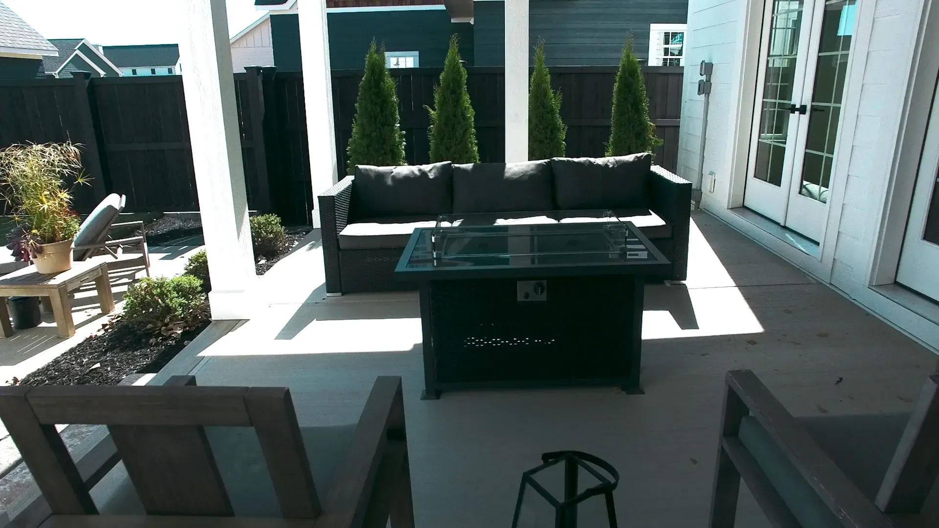 Patio with gray couch, fire pit, and chairs under a white-columned porch, with green trees in the background.