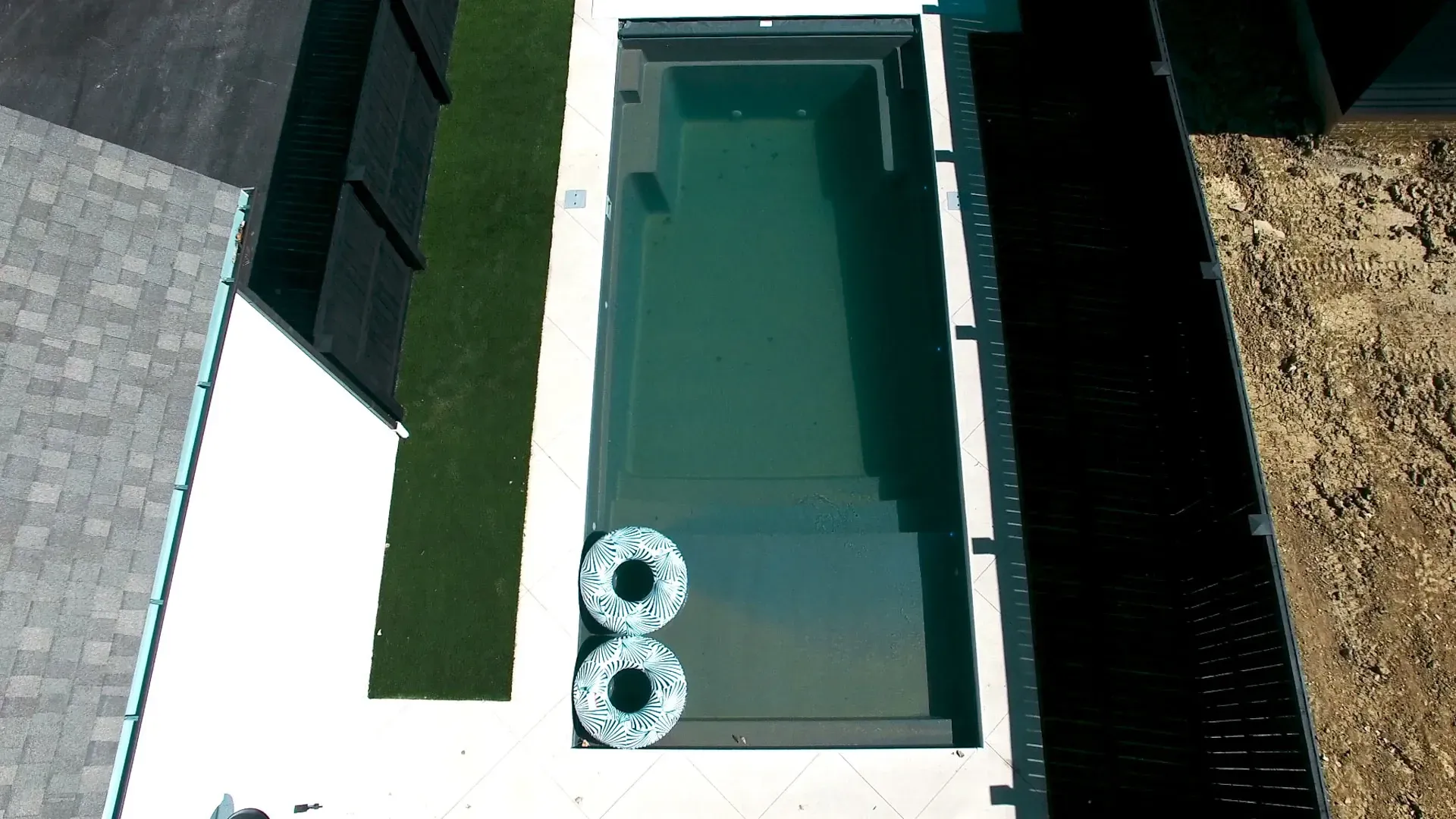 Aerial view of a rectangular swimming pool with two floating rings. The pool is surrounded by concrete and green lawn, next to a building.