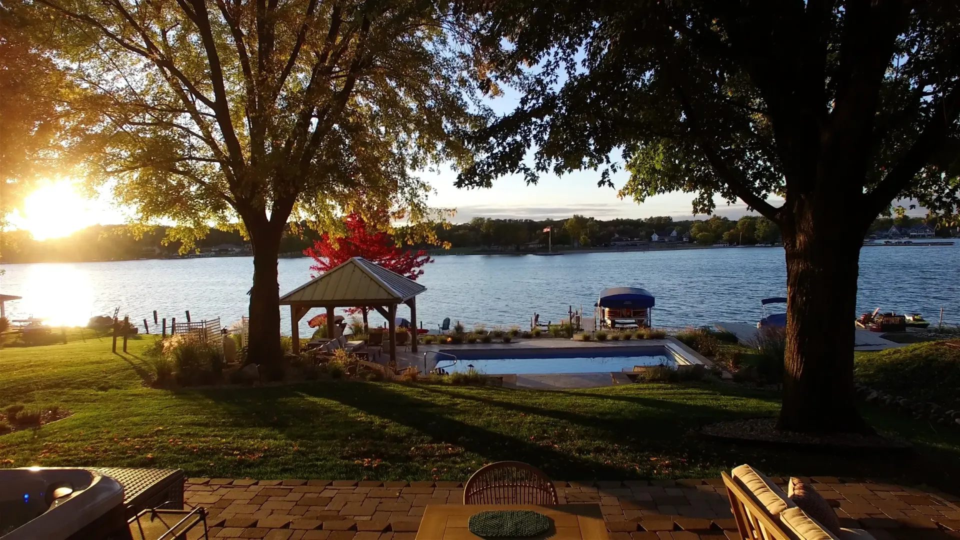 Lakeside backyard scene at sunset with a pool, gazebo, and trees. Sun reflects on the water.