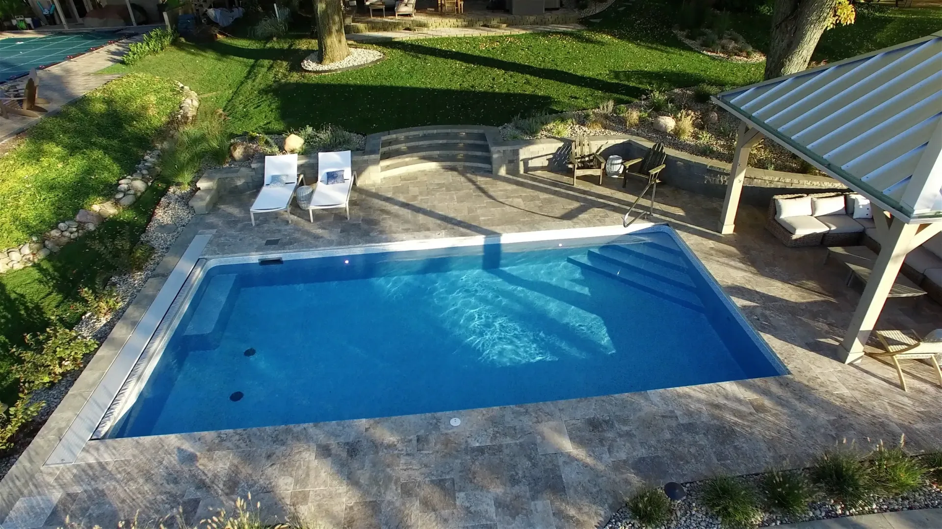 A rectangular swimming pool with blue water surrounded by a stone patio, lounge chairs, and a gazebo.